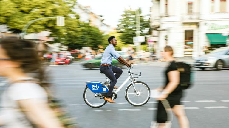 Imagen de una escena callejera con peatones y un ciclista. Al fondo se ven árboles.